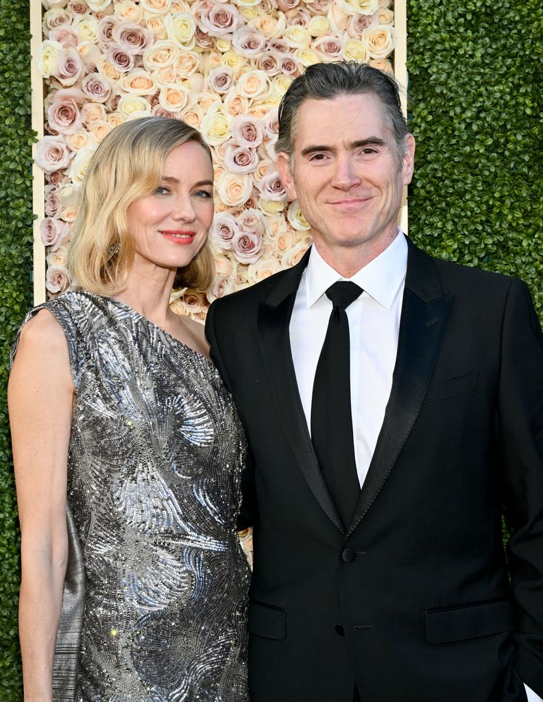 Naomi Watts in a silver dress with Billy Crudup against a backdrop of flowers