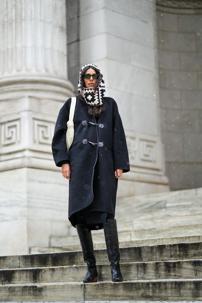 Babba wears oversized black duffle coat with statement buttons and her crochet balaclava adds a modern twist. The tall black boots are functional and chic, m