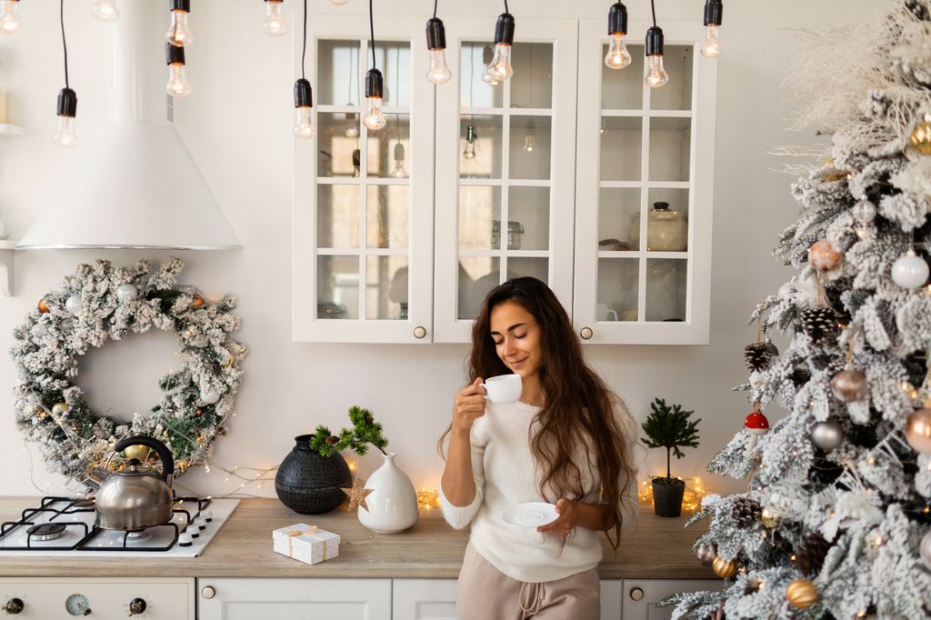 Young woman making hot tea at home and prepare Christmas meal. Home is decorated with Christmas tree, ornaments, wreath and garland lights.