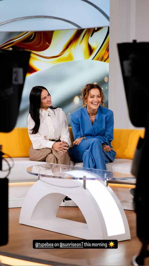 Rita Ora and co-founder Anna Lahey share laughs on a morning talk show, dressed in sleek suits with minimalist styling.