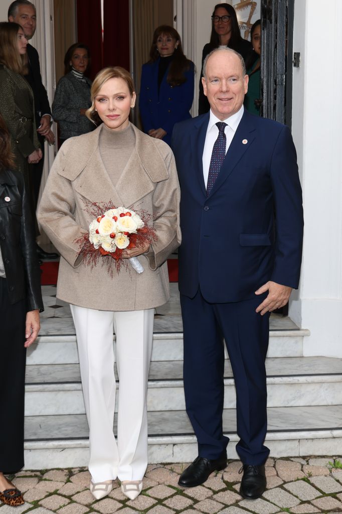 Prince Albert in suit and Princess Charlene in white trousers and taupe coat holding flowers