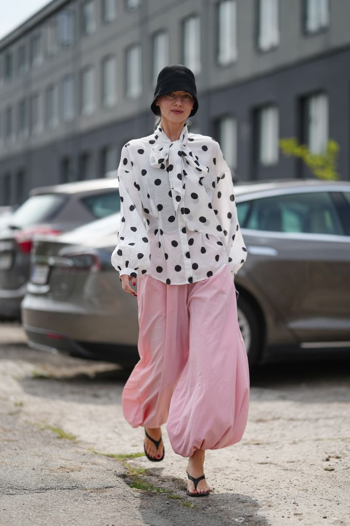A guest wears a black bucket hat with light blonde hair tucked underneath, soft pink lipstick, a white lightweight woven blouse with oversized black polka dots, a self-tie bow at the neck, and billowy long sleeves with gathered cuffs, paired with voluminous pale pink balloon-hem trousers in a flowy fabric gathered at the ankles, black flat flip-flops with thin straps, and bright red nail polish on fingernails, walking, outside Marimekko, during Copenhagen Fashion Week,