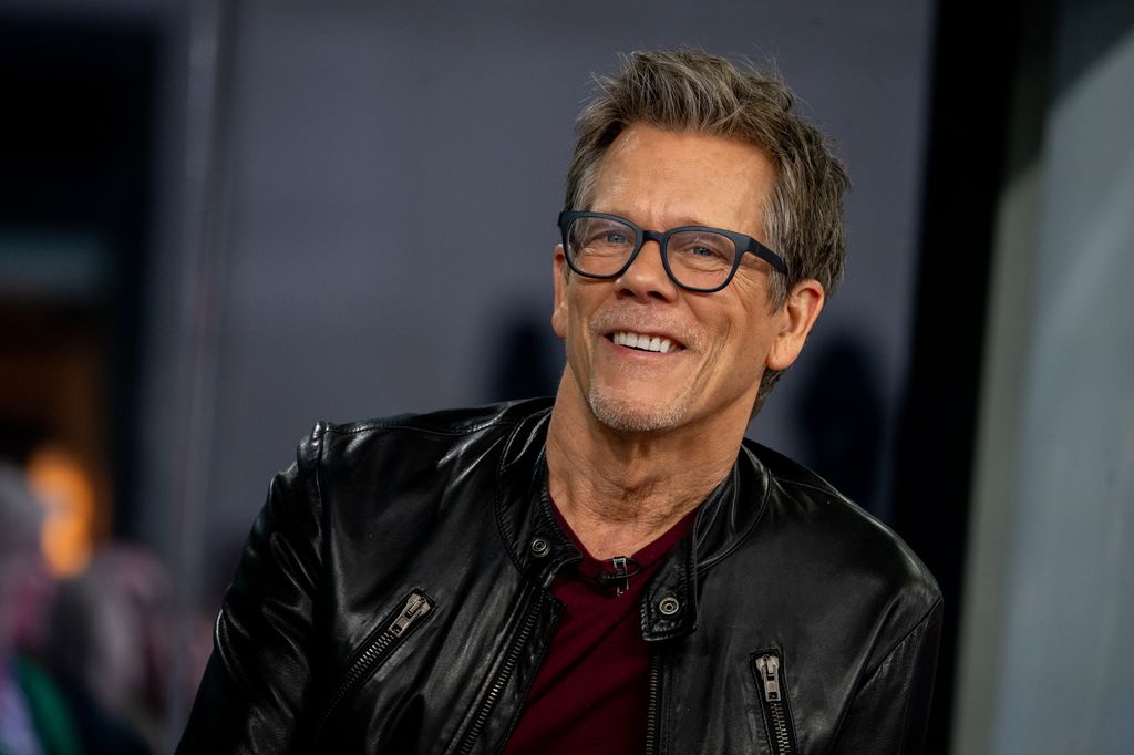 Kevin Bacon smiles wearing leather jacket and glasses