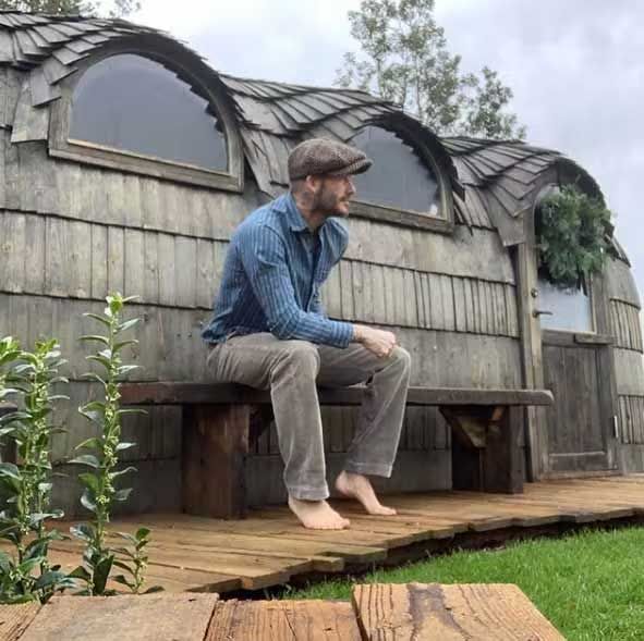 david wears a flat cap and sits outside on a bench in front of large wooden huts with semi circle windows