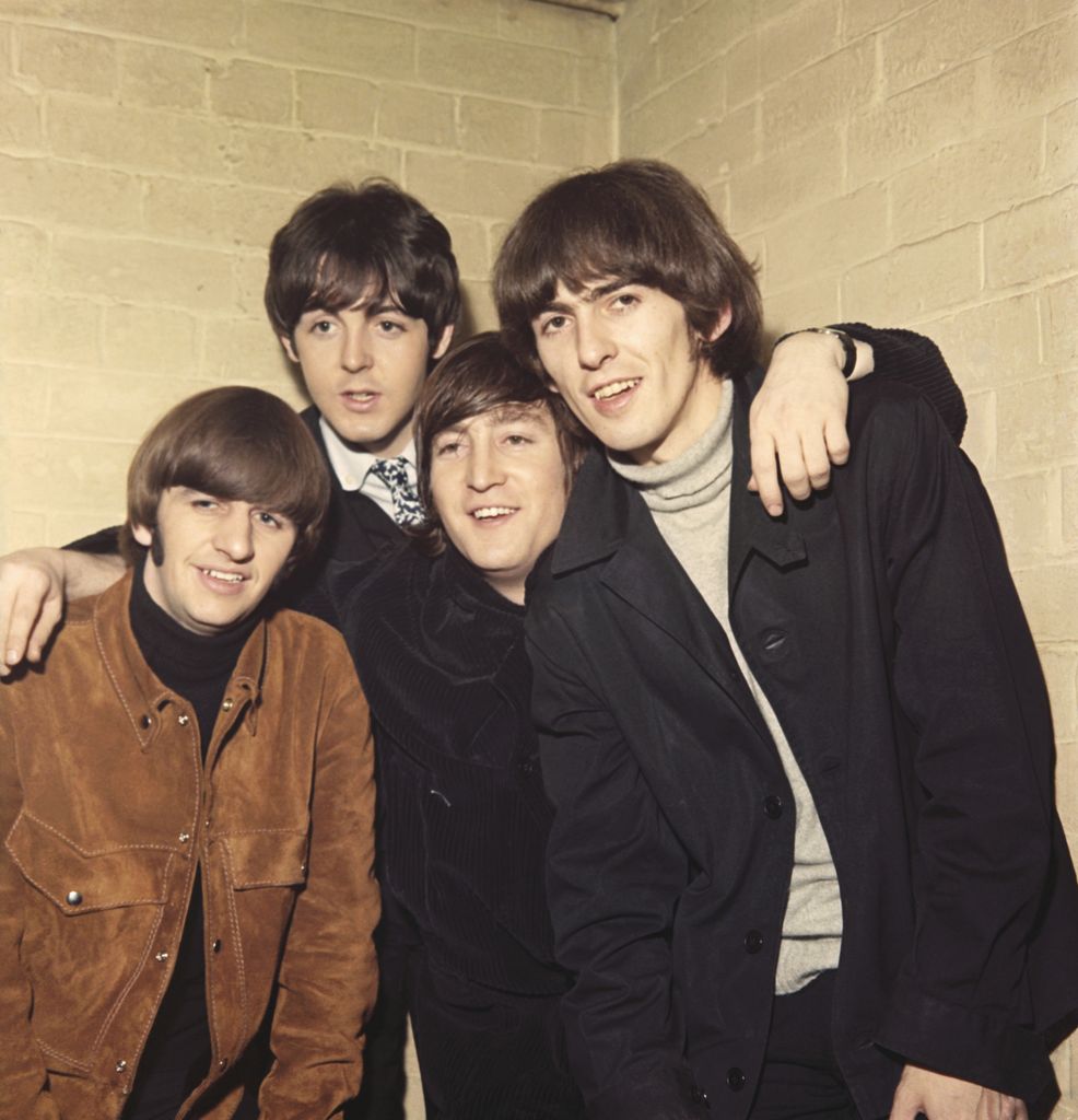 The Beatles huddle together, grinning in warm jackets and mod-style turtlenecks. They're young, fresh-faced, and look like they just finished recording the coolest song of 1965.