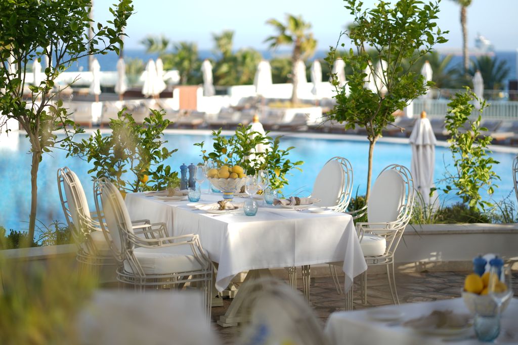 utdoor dining table set with white tablecloth, cutlery, and lemons, surrounded by white chairs and greenery, overlooking a pool with loungers and umbrellas in the background.