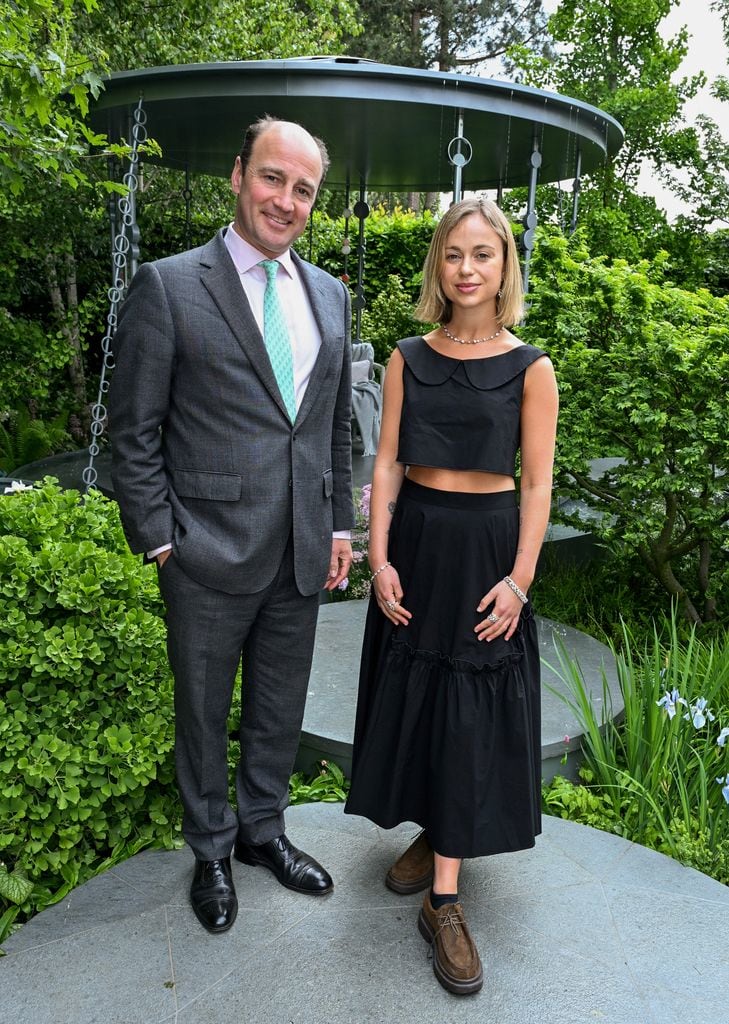James Amos in a grey suit stands next to Lady Amelia Windsor in a black sleeveless top and matching skirt with brown shoes at Chelsea Flower Show 2025.