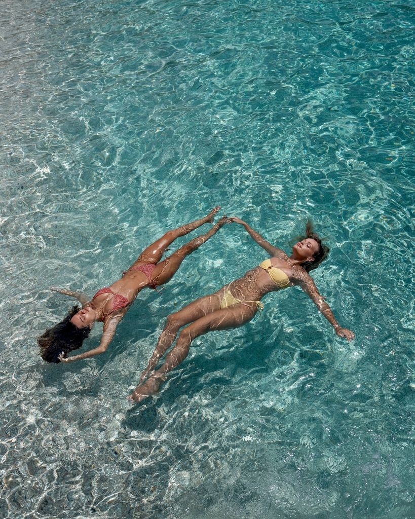 Two models float in crystal clear blue ocean water wearing bikinis