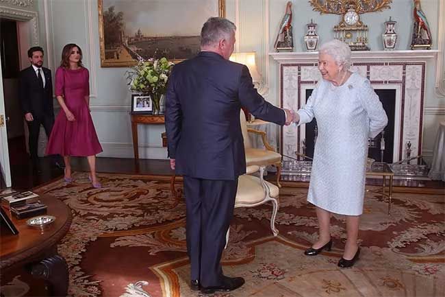 the queen wears a pale blue dress as she stands shaking hands with a navy blue suited man in a room with burgundy and gold carpet a fireplace in the background and white walls decorated with gold framed paintings
