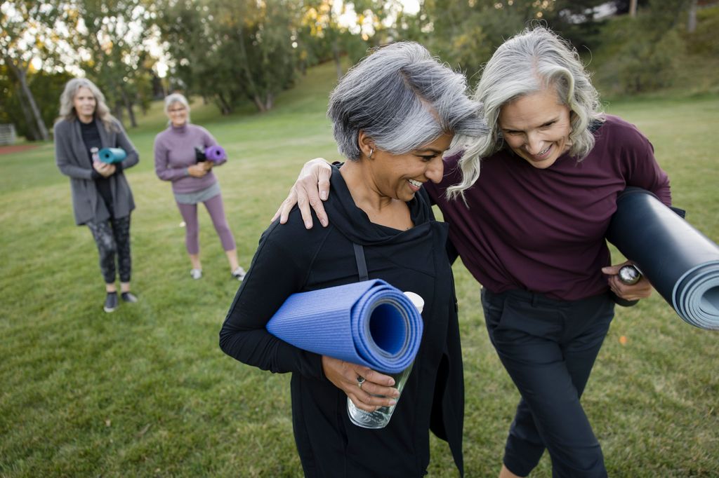 Happy mature women friends hugging, walking and laughing with yoga mats in park