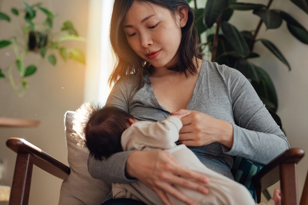Young Asian mother breastfeeding her baby daughter while sitting on sofa at home