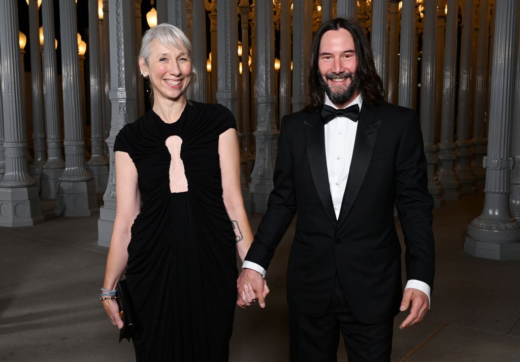 Alexandra Grant and Keanu Reeves smiling and holding hands in black outfits