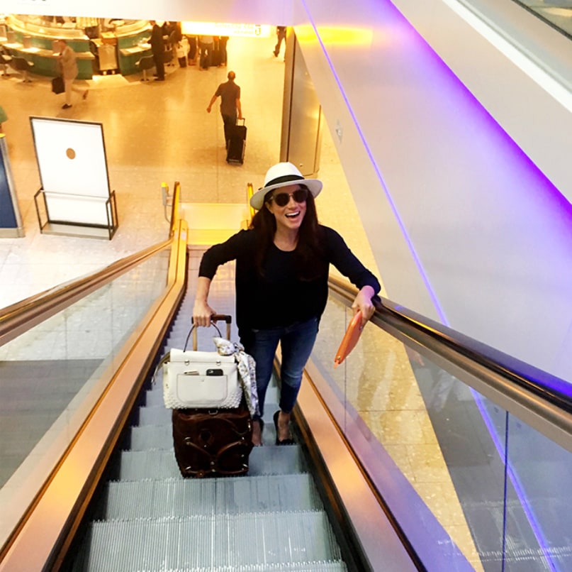 Meghan Markle wears sunglasses and a Panama hat, holding a suitcase on an escalator at the airport