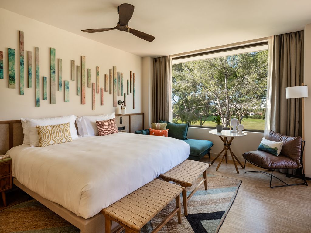 Elegant guest room at Kimpton Aysla Mallorca featuring a plush bed with decorative pillows, unique wooden wall art, and a bright window view of verdant gardens.