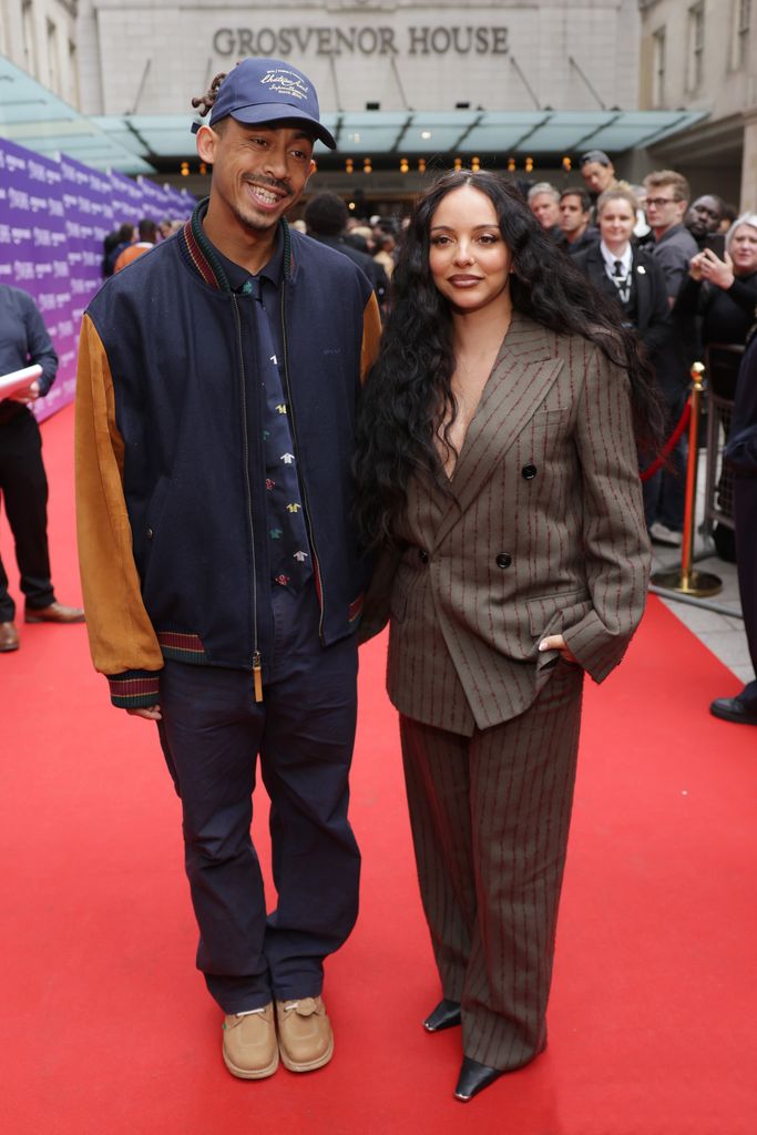 Jade Thirlwall and Jordan Stephens stand on the red carpet, Jade is wearing a brown pinstripe suit with black heels and Jordan is wearing jeans, a baseball jacket and cap and beige boots