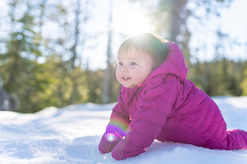 An adorable one year old baby girl wearing a pink snowsuit smiles while crawling in the snow