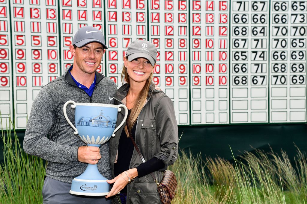 Rory McIlroy holding a blue trophy alongside Erica Stoll in front of a scoreboard