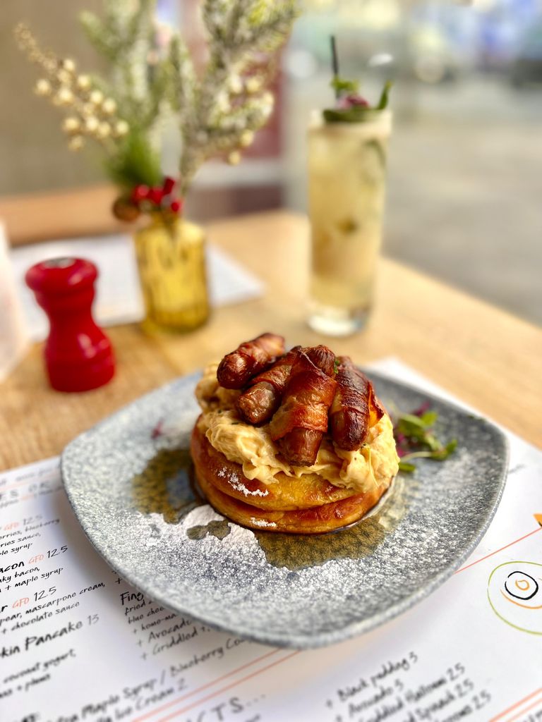 A festive Christmas brunch dish from The Table restaurant, featuring fluffy pancakes topped with creamy scrambled eggs and crispy bacon-wrapped sausages, served on a decorative plate.