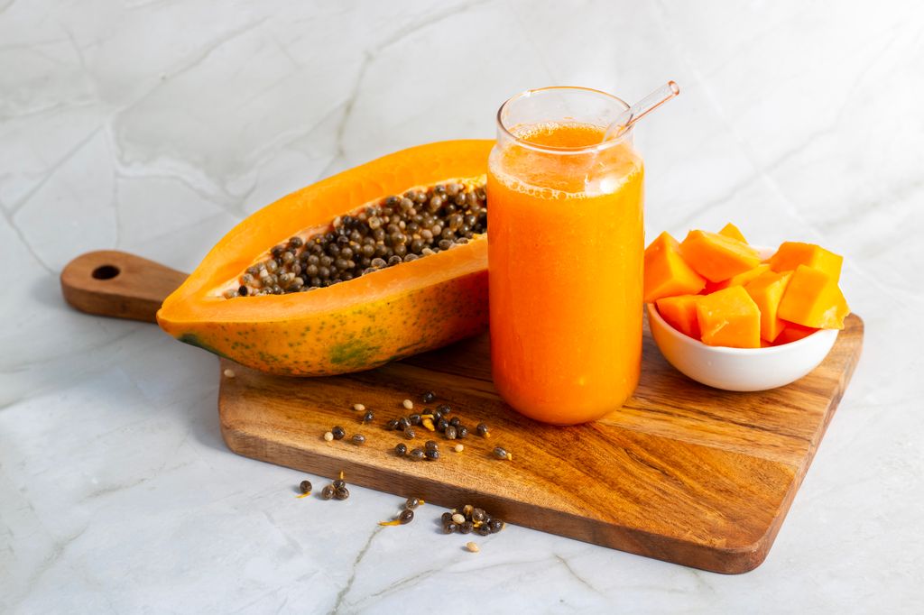 A halved papaya, vibrant smoothie in a glass jar with a straw, fresh cubes in a bowl, and scattered seeds on a wooden board.