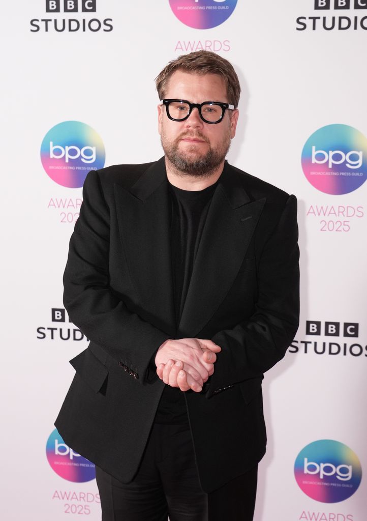 James Corden in a black suit clasping his hands together while standing in front of BBC Studios step and repeat board