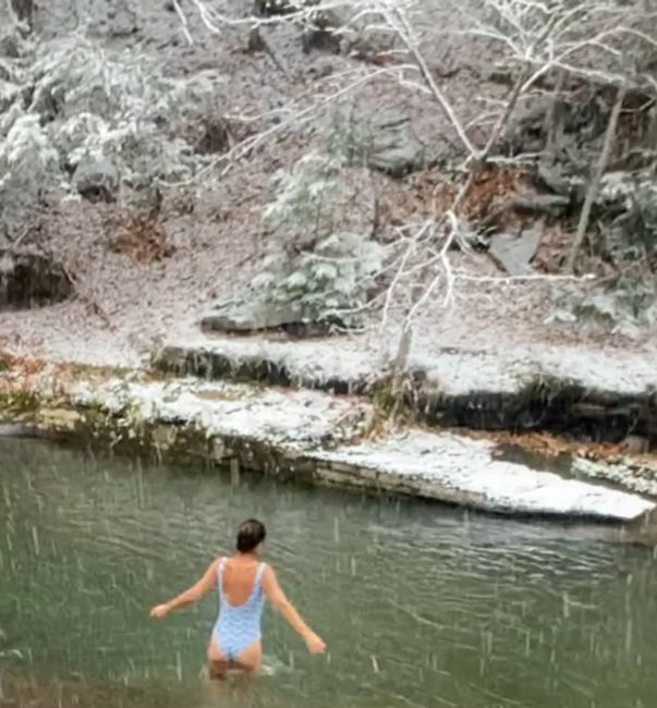 a wide shot of helena wearing a low back swimsuit as she wades into hip deep water as snowfalls around her and on the riverbank