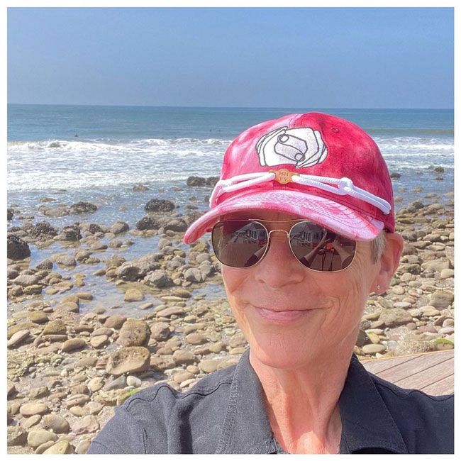 jamie lee curtis takes a selfie on the beach wearing a red cap