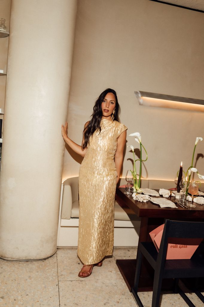 Jasmin Larian Hekmat poses in a crinkled gold floor-length gown beside a candlelit dinner table adorned with calla lilies at The Holy Carrot, hosting an intimate celebration for Cult Gaia’s latest collection in Notting Hill.