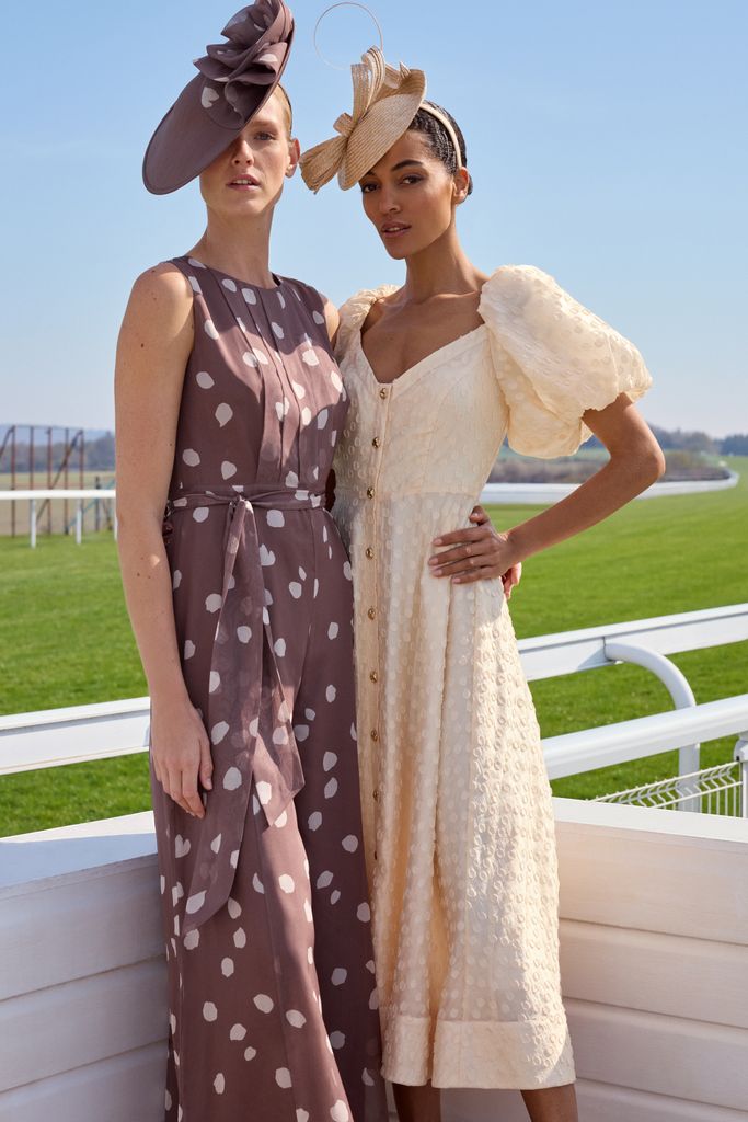 Two women standing in front of a race track, one wears a brown polka dot dress and matching fascinator, the other wears a puff sleeved midi dress and small headpiece