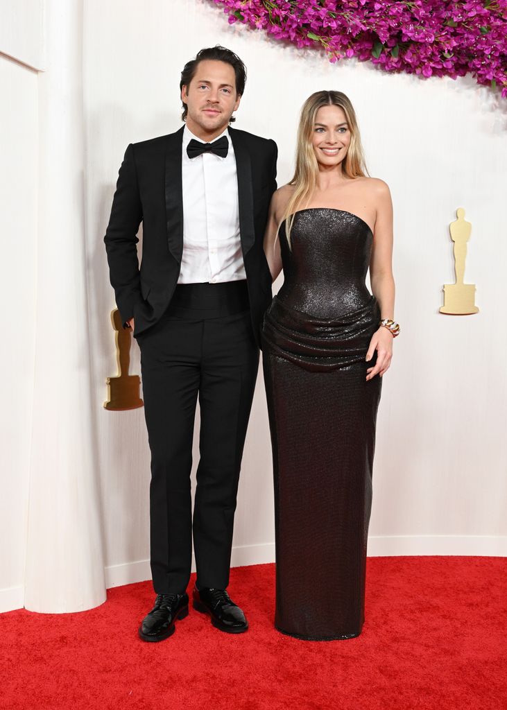 Tom Ackerley and Margot Robbie smile for photographers in front of the white screen while visiting Oscars