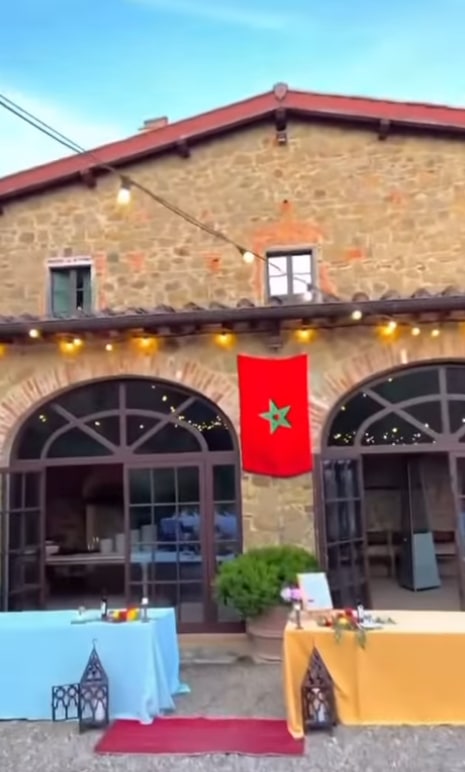 A rustic building with arched windows is decorated with string lights and a Moroccan flag. Two tables covered in blue and yellow cloth are set up outside, with lanterns and greenery nearby under a clear sky.