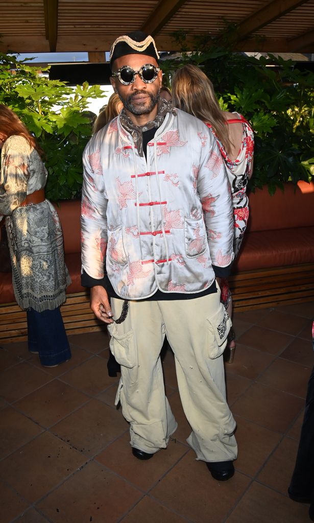Terence Eben wears a silk embroidered jacket with crane motifs, beige cargo trousers, steampunk sunglasses and a velvet hat at Chelsea Flower Show 2025