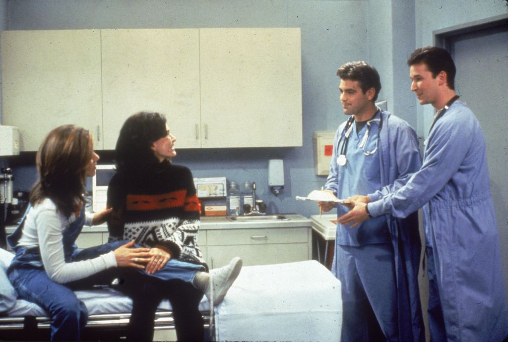 Jennifer Aniston and Courteney Cox sit in a hospital room, speaking to guest stars George Clooney and Noah Wyle from 'ER' in a still from the television series, 'Friends,' circa 1996
