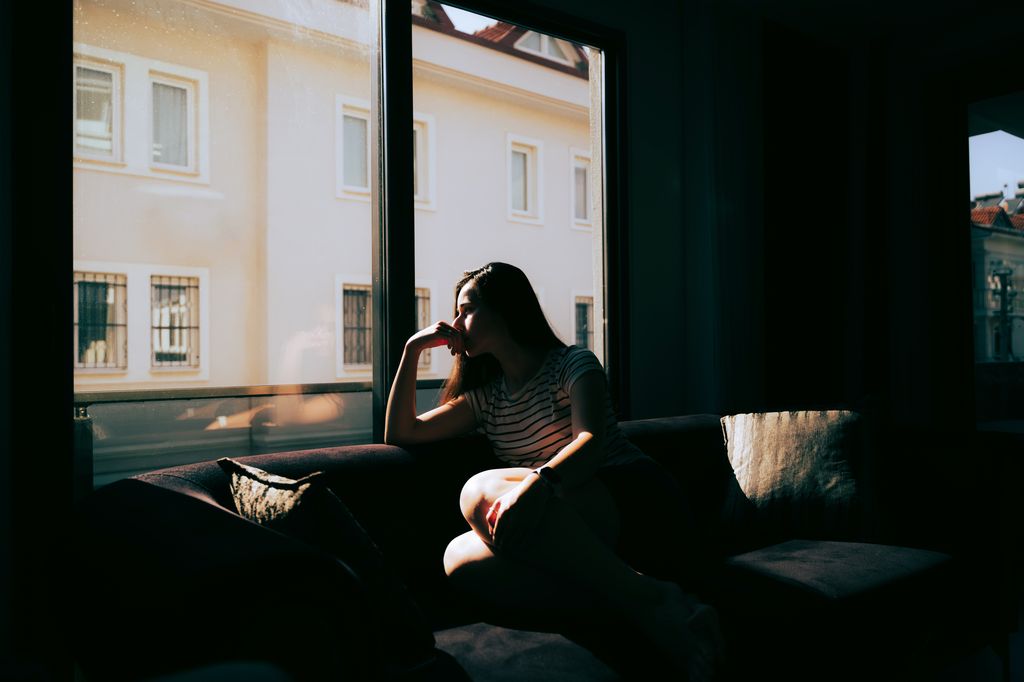 A tired and depressed  woman is sitting on the sofa near the window at home