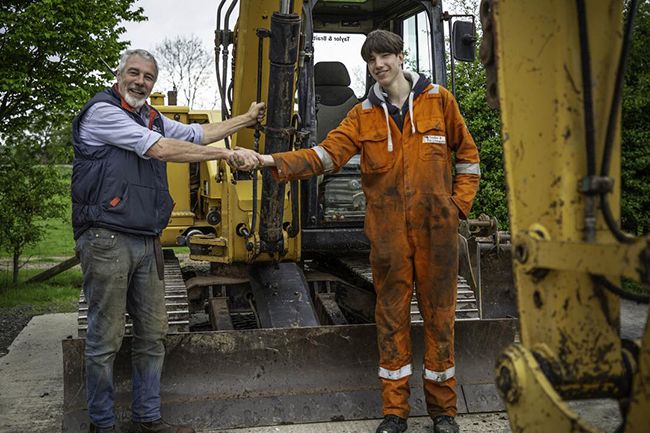 Reuben and Clive Owen shakes hands in front of digger