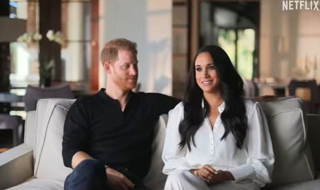 harry sits on a sofa next to meghan who stands out wearing an all white ensemble consisting of a shirt and trousers