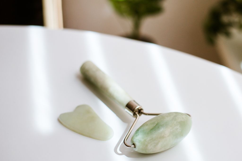 Close-up gua sha scraper and roller on white table with sunbeams with plants on background.