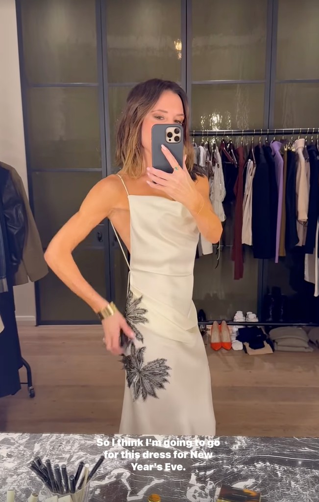 Victoria Beckham poses in her dressing room wearing a cream cowl neck slip dress with black embroidered detailing on the side and shoulder. She teams the dress with a gold cuff and stands in front of her makeup station, her hair coiffed.