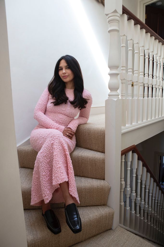 Nieve Tierney sits on a beige carpeted staircase wearing a long-sleeved pink textured knit dress and black leather loafers, with relaxed hair and a serene expression.