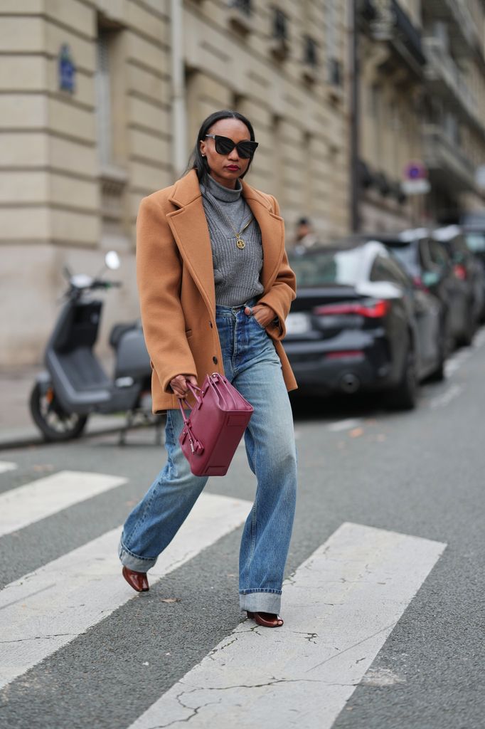 Emilie Joseph wears long straight dark brown hair parted at the center, makeup featuring an even complexion with matte skin and deep red lipstick, oversized black sunglasses by Gentle Monster, layered knitwear consisting of a light grey wool turtleneck under a slouchy knit pullover by Patou, a camel sculptural wool peacoat with strong shoulders and a couture martingale detail by Saint Laurent, blue V-waist high-rise denim jeans pants by Saint Laurent, a burgundy leather handbag by Saint Laurent YSL, and chestnut patent leather peep-toe platform sandals by Saint Laurent, during a street style fashion photo session on December 13, 2025 in Paris, France. (Photo by Edward Berthelot/Getty Images)