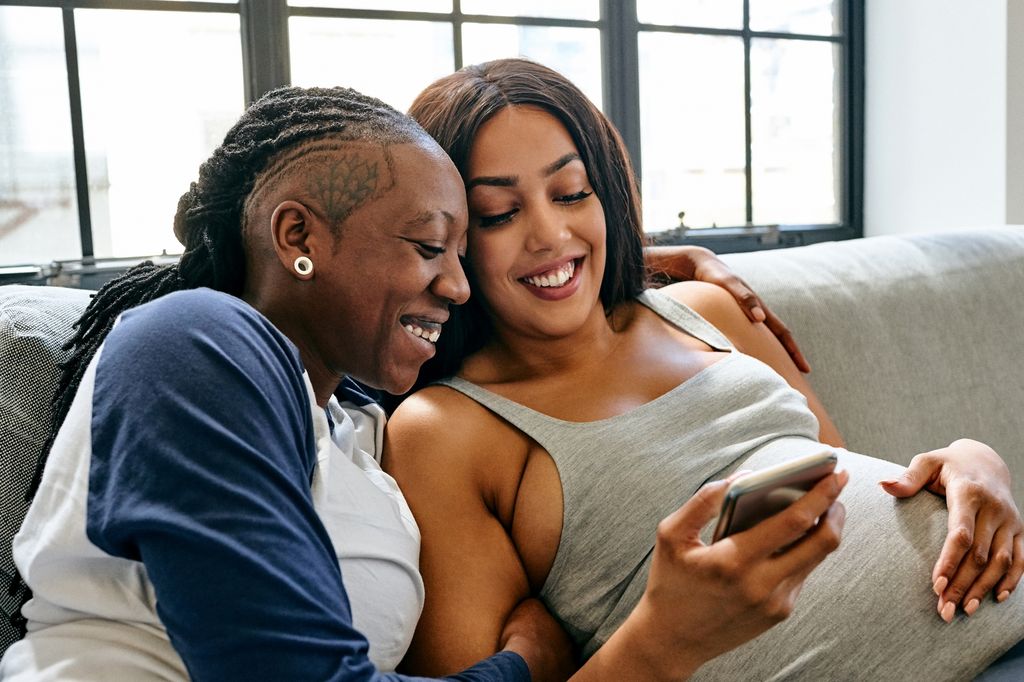 Lesbian couple looking at mobile phone and smiling in living room at home.