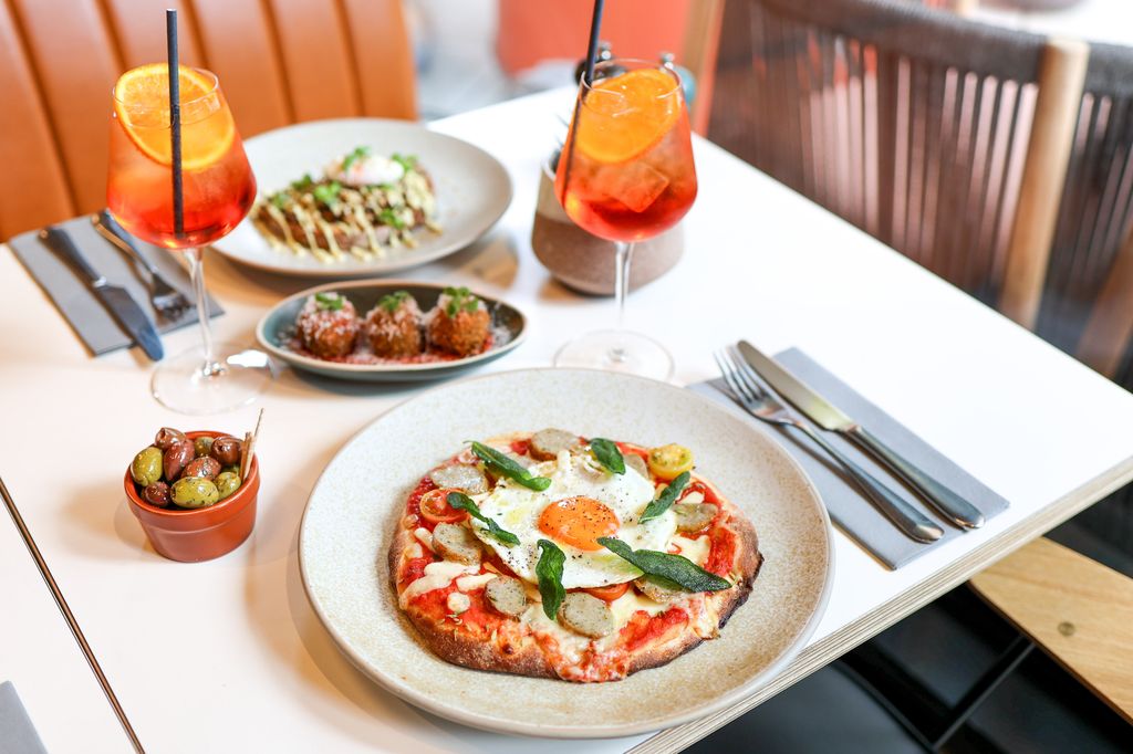 table with pizza and arancina and pot of olives, with two wine glasses of aperol spritz