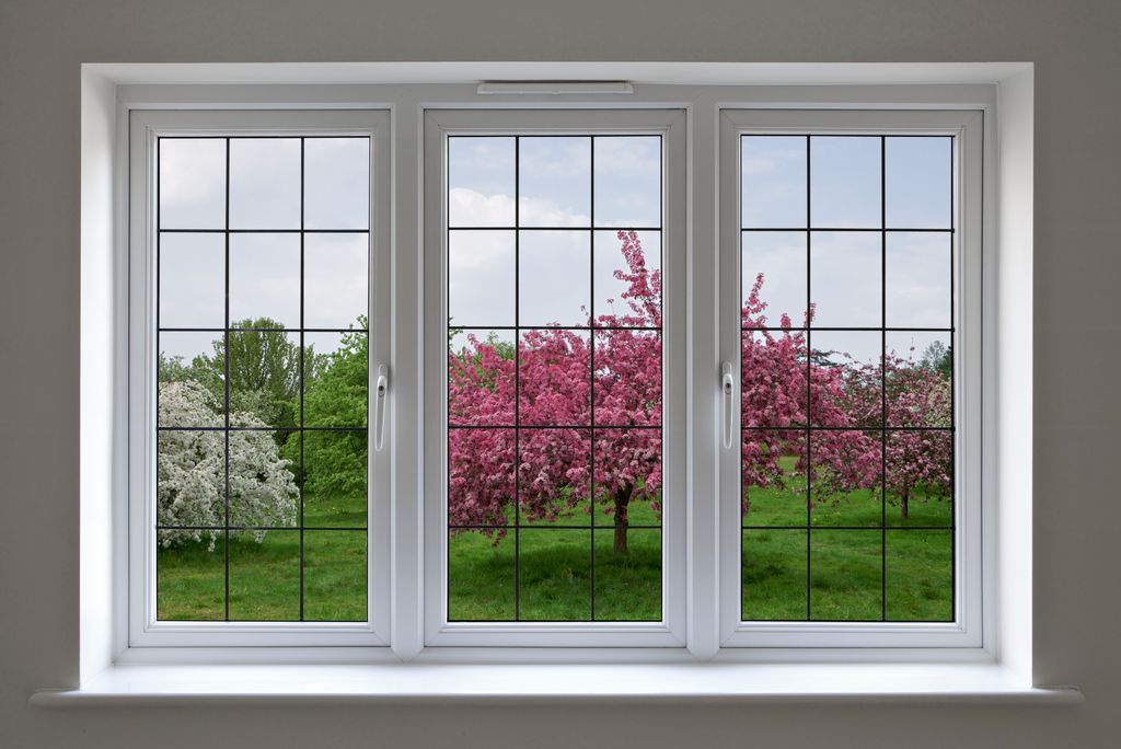 Beautifully made white wood framed windows with leaded glass panels, surrounded by a light grey wall.  The view through the window is over an apple orchard under a cloudy blue sky. This is a composite of two images.