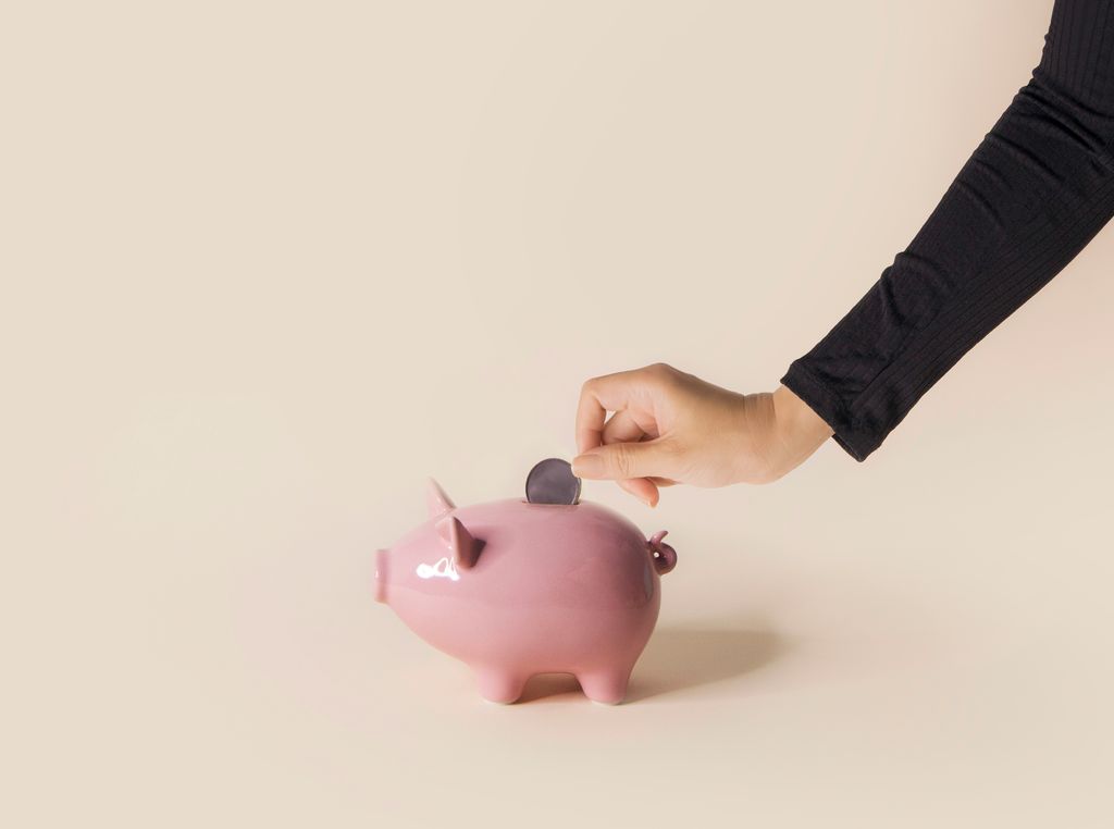 Hand putting coin into piggy bank on cream coloured background.