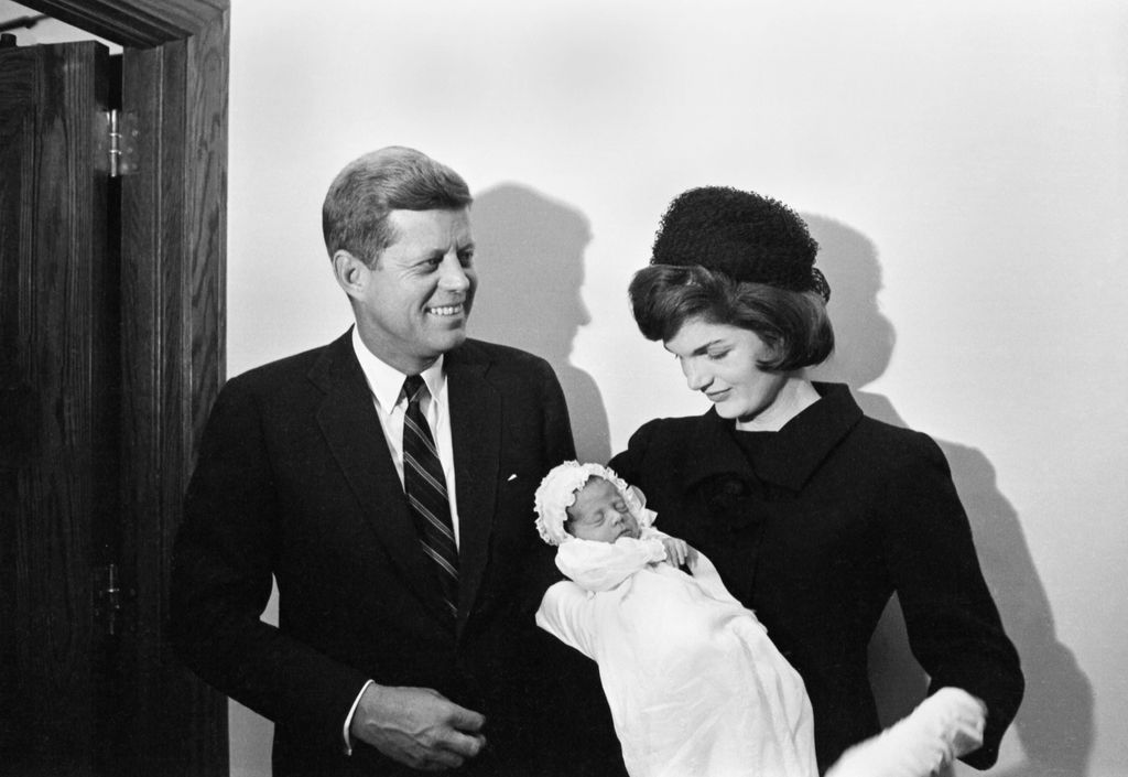 John Fitzgerald Kennedy, Jr., is photographed for the first time at the age of 13 days during his christening in the Georgetown University Hospital Chapel here, December 8th. The infant is being held by his mother, Mrs. Jacqueline Kennedy. The proud papa, President-elect John F. Kennedy, smiles beside her.