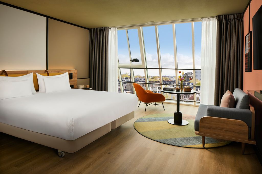 Modern hotel room in Amsterdam with a large bed, floor-to-ceiling windows, city view, and stylish seating area
