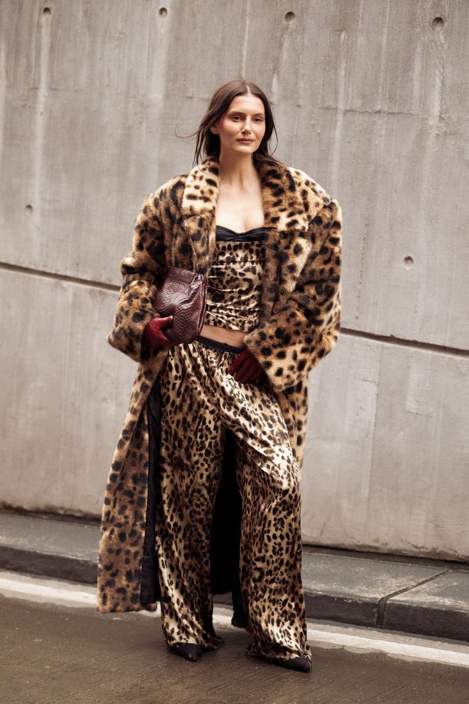  guest wears a leopard-print faux fur coat over a matching leopard set, styled with red leather gloves and carrying a brown textured clutch outside the OperaSport show