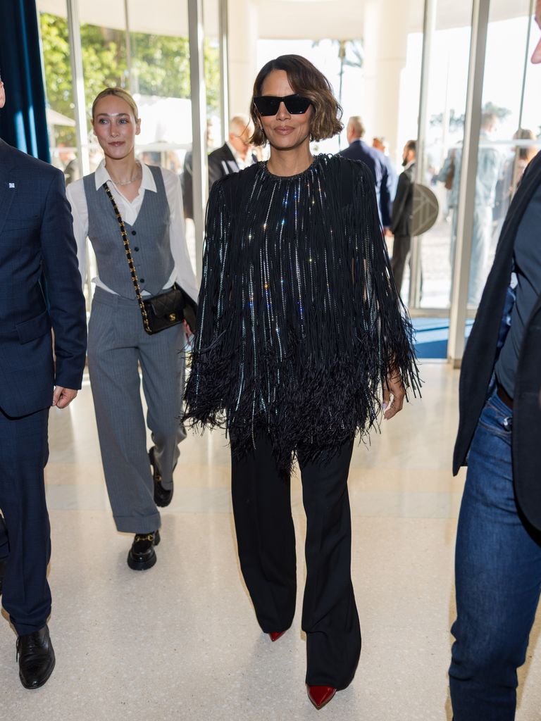 Halle Berry wears black sunglasses, a black metallic fringed cape, black flared trousers and deep red pointed heels. 