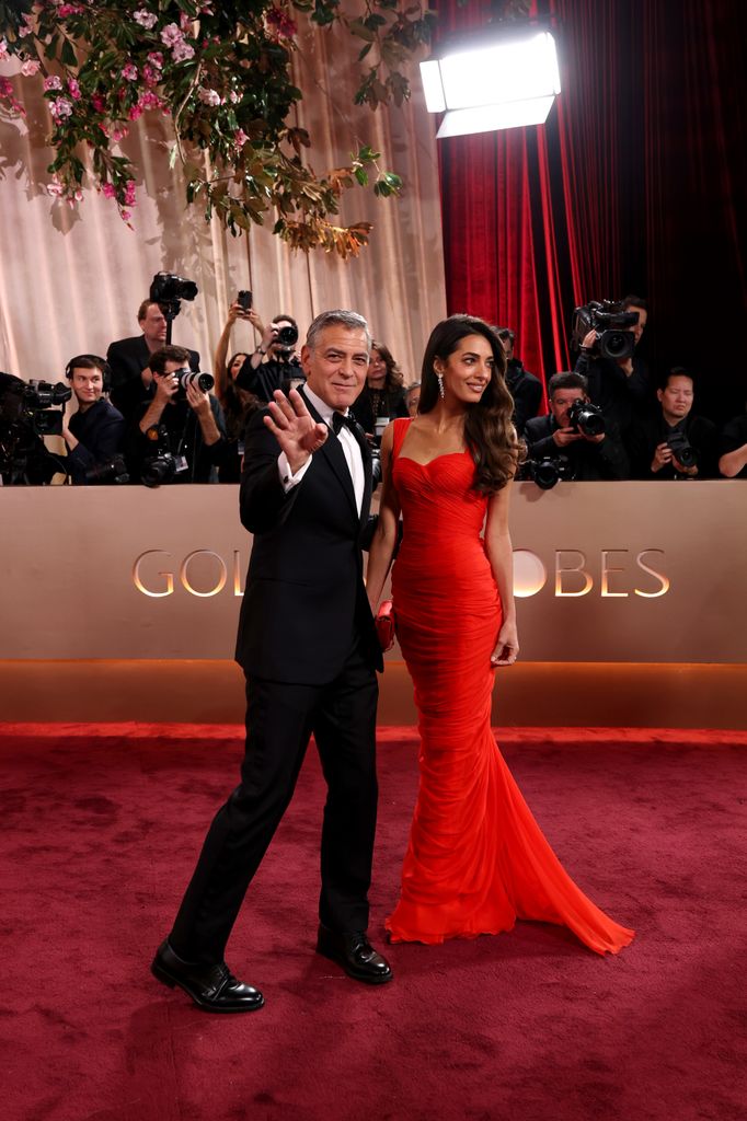 George Clooney and Amal Clooney walk the red carpet, Amal in a red dress and George in a black suit and white shirt