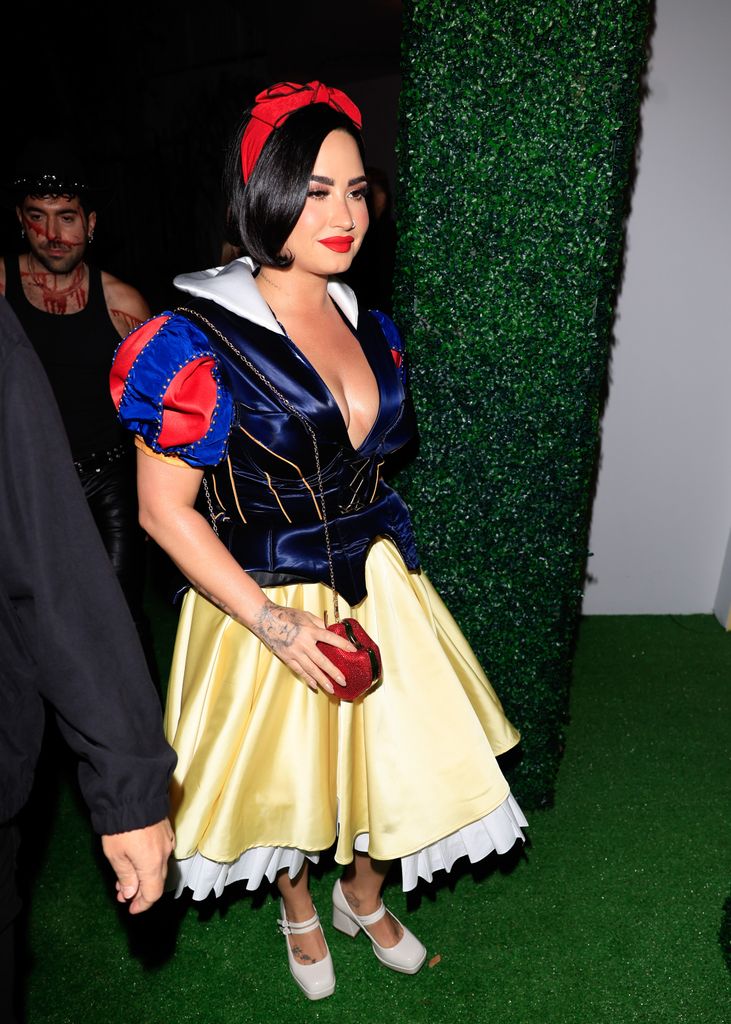 Demi Lovato  dressed as Snow White in a fitted blue and yellow gown with a red headband. They hold a red apple-shaped purse, and their black hair is styled short and sleek.