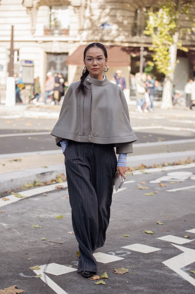A guest wears a light grey structured cape jacket layered over a blue striped shirt and pinstripe wide-leg trousers, accessorised with hoop earrings and clear glasses outside Hermes show during the Womenswear Spring Summer 2026 as part of Paris Fashion Week 
